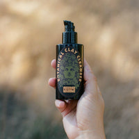 A hand holding a black pump bottle of Botanical Clay Cleanser against a blurred outdoor background.
