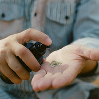 A close-up of a person's hands holding a small droplet of white cream or serum in their palm while spraying a dark bottle, with a striped shirt in the background.