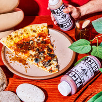 A hand holds a squeeze bottle of Mexican Barbecue sauce next to a slice of pizza on a plate, with another bottle lying on an orange textured cloth and more food props around it.