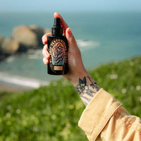 A hand holding a black spray bottlE against a blurred outdoor background of green grass and the ocean.