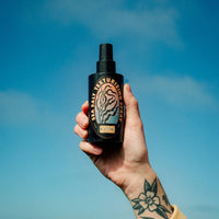 A hand with a floral tattoo holds up a black spray bottle of hair texture spray against a bright blue sky background. The bottle features a black label with a stylized tiger graphic and gold text reading 'Sea Salt TEXTURE SPRAY' and 'CHURCH'.