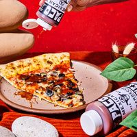A hand squeezes a bottle onto a slice of pizza on a plate, with another bottle lying nearby on a rustic red and orange styled table.