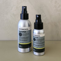Two silver spray bottles of Hydrosol Juniper by All Spas on a beige surface against a textured beige wall.