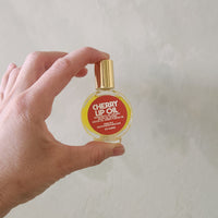 A hand holding a small round bottle of cherry lip oil with a gold cap against a textured off-white wall.