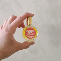 A hand holding a small round glass bottle of Strawberry Lip Oil with a gold cap against a textured light gray background.