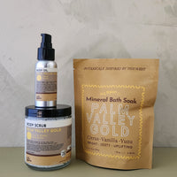 A bath soak product set including a gold pouch labeled 'Mineral Bath Soak Palm Valley Gold,' a silver pump bottle labeled 'Body Oil Palm Valley Gold,' and a clear jar labeled 'Body Scrub Palm Valley Gold,' arranged on a tabletop against a textured light gray wall.