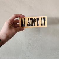 A hand holds a rectangular beige sticker or card with the phrase 'AI AIN'T IT' in bold black letters against a textured off-white wall.