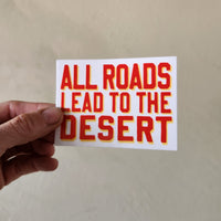 Hand holding a sticker with 'All roads lead to the desert' text on a plain background