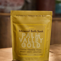 A stand-up pouch of Palm Valley Gold Mineral Bath Soak by All Roads, featuring a golden-yellow design with black and white text, set on a wooden surface with a blurred rustic background.