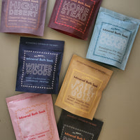 Seven pouches of botanical-inspired mineral bath soaks in assorted scents, arranged in a flat-lay pattern on a neutral beige background. The pouches come in a variety of colors including purple, burgundy, light blue, navy, mustard yellow, pink, and black, each with its own unique illustrated label.