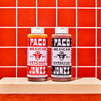 Two bottles of taco seasoning from Paco Jones Mexican Ketchup and Barbecue Seasoning displayed side-by-side on a light wood block against a red tiled background.