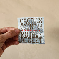Hand holding a sticker with 'Cactus Country Mojave Desert' text against a beige background