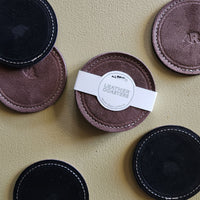 Six round leather coasters in black and muted mauve-brown tones arranged on a textured beige surface. One coaster has a white paper band labeled LEATHER COASTERS' wrapped around it.
