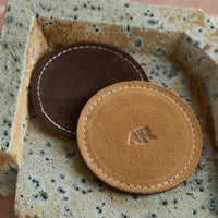 Two leather coasters with a visible brand logo on a textured surface
