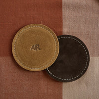 Two leather coasters with 'AR' logo on a checkered fabric background