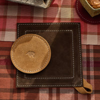 Brown leather potholder with tan coaster on a red and brown checkered tablecloth.
