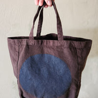 Brown tote bag with a blue circular design held by a hand against a neutral background