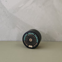 A single cylindrical cosmetic container lies on its side on a muted sage surface against a light gray textured wall. The product label reads 'COASTAL CREME POWDER' and appears black with teal and gold text.