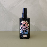 A black spray bottle of Church Sea Salt Texturizing Spray with a colorful label, set against a textured light gray wall and a beige surface.