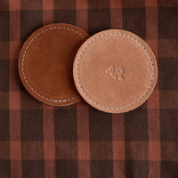 Two brown leather coasters with a logo on a plaid fabric background