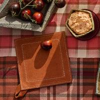 Brown leather coaster with a tomato on a red and beige checkered tablecloth.