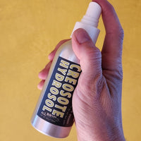 Hand holding a bottle labeled 'Creosote Hydrosol' against a yellow background