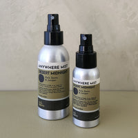 Two silver spray bottles of 'Anywhere Mist' in 'Desert Midnight' scent, one larger and one smaller, standing on a beige surface against a textured beige wall.