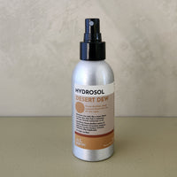 A bottle of Hydrosol Desert Dew room spray with a silver metallic body, black spray nozzle, and a beige and brown label, set against a textured beige background.