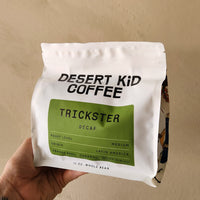 Hand holding a 'Desert Kid Coffee' package labeled 'Trickster Decaf' against a beige background