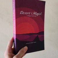 Book titled 'Desert Magick' held by a hand against a neutral background