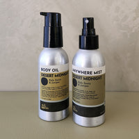 Two bottles of All Roads body and fragrance products, 'Body Oil' and 'Anywhere Mist', in silver metal bottles with black pumps and black-and-beige labels, set against a neutral beige background.