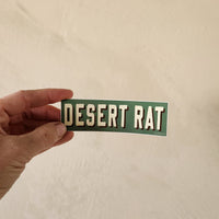 Hand holding a 'DESERT RAT' sticker against a beige background