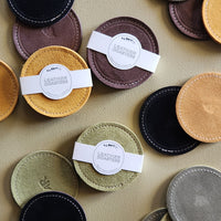 A flat-lay arrangement of circular leather coasters in multiple colors, including black, brown, mustard yellow, sage green, olive, tan, and gray, each with white stitched edges and a white paper band label reading 'LEATHER COASTERS'