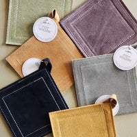Six square suede-like pouches or coasters in assorted muted colors, arranged in a flat lay on a beige surface. Each item has a small loop tab, a round white hangtag reading 'POTHOLDERS', and visible contrast stitching along the edges.