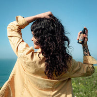A woman with long, curly black hair is shown from behind outdoors, holding a small black spray bottle to her hair. She is wearing a light beige jacket, with a blue sky and green grass in the background.