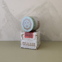 A jar of Goldie Goldie hair pomade resting on top of its branded cardboard box, set against a textured light gray wall and a muted olive-green surface.