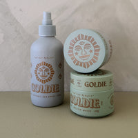 Three Goldie brand hair care containers on a beige surface against a textured beige wall: a matte spray bottle, a round jar, and a smaller round tin stacked on top. The bottle is light gray with tan sun graphics, while the jars are pale mint green with tan sun graphics.