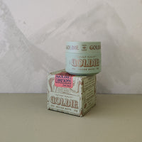 A small jar of Goldie Mellow Matte hair pomade sits on top of its matching printed box, photographed against a light textured wall and neutral tabletop.