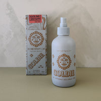 A bottle of Goldie Sea Spray next to its retail box, both featuring a gold sun face logo on a white background, set against a textured beige wall and surface.