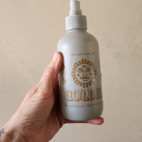 A hand holding a frosted white spray bottle labeled 'GOLDIE' with a gold sun face logo, against a plain beige wall.