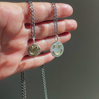 Two smiley face pendants on chains held in a hand against a neutral background