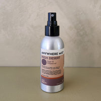 A silver spray bottle of 'Anywhere Mist' in 'High Desert' scent, standing on a beige surface against a textured beige wall.