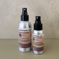 Two silver spray bottles of Anywhere Mist body mist, one labeled High Desert and one labeled Desert Midnight, standing on a beige surface against a textured beige wall.