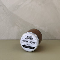 A glass jar of H.W. Jones Hair Mask lies on its side on a textured beige surface against a neutral gray wall.