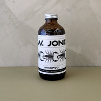 An amber glass bottle of W. Jones Shampoo with a white label and silver cap, set against a neutral textured beige background.