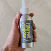 Hand holding a Juniper Hydrosol bottle against a neutral background