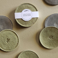 A set of round leather coasters arranged on a textured beige surface. One central coaster has a white paper band wrapped around it with a circular label that reads 'LEATHER COASTERS'
