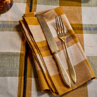A folded linen napkin in a mustard yellow, beige, and brown plaid pattern, styled with a vintage-style knife and fork on a matching plaid tablecloth.