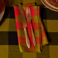 A folded red and olive green plaid napkin with a vintage-style gold knife and fork, set on a matching plaid tablecloth.