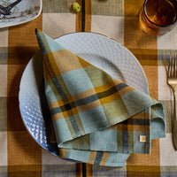 A folded plaid napkin in muted sage green, mustard yellow, and navy is arranged on a white plate at a table setting with a matching plaid tablecloth, gold-toned cutlery, and a small decorative dish.